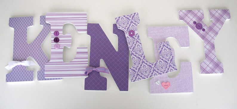 May include: Purple and white fabric letters spelling the name "KENLY" with decorative buttons and bows. The letters are made of wood and have a 3D effect.