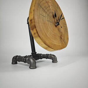 May include: A unique wooden clock with a natural wood grain face. The clock is supported by a black metal pipe base. The clock hands are black. The clock face is approximately 10 inches in diameter.