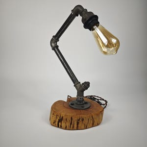 Puede incluir: Lámpara de escritorio de estilo industrial con una bombilla Edison vintage. La lámpara tiene una estructura de tubería metálica y una base de madera. La bombilla es de color ámbar cálido. El diseño recuerda la estética steampunk.