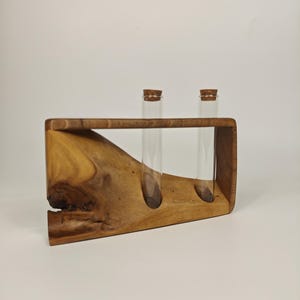 Könnte beinhalten: Ein Holzständer für Reagenzgläser mit zwei Glasröhrchen, die jeweils mit einem Korken verschlossen sind. Der Ständer besteht aus hellbraunem Holz mit natürlicher Maserung und organischer Form. Die Röhrchen sind transparent und leer.