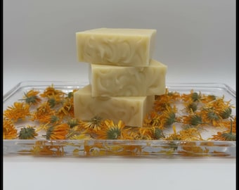 Könnte beinhalten: Vier hellfarbene Seifenstücke, gestapelt auf einem durchsichtigen Tablett, umgeben von getrockneten orangefarbenen Ringelblumenblüten. Die Seife hat ein Wirbeldesign auf der Oberseite. Der Hintergrund ist schlicht weiß.