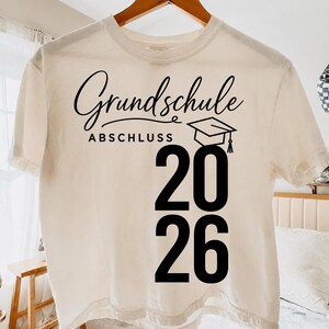 May include: A cream-coloured short-sleeve t-shirt featuring black text that reads Grundschule Abschluss with a graduation cap icon and the large numbers 20 26 printed vertically on the right side of the garment.