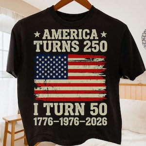 Puede incluir: Camiseta negra con el texto "AMERICA TURNS 250" sobre una bandera americana desgastada. Debajo, se lee "I TURN 50 1776-1976-2026". El dise&ntilde;o utiliza blanco, rojo y azul. La camiseta est&aacute; colgada en una percha de madera.