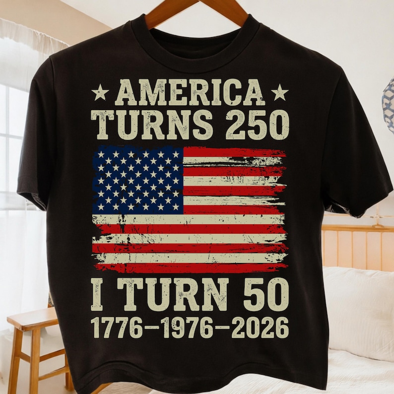 Puede incluir: Camiseta negra con el texto "AMERICA TURNS 250" sobre una bandera americana desgastada. Debajo, se lee "I TURN 50 1776-1976-2026". El dise&ntilde;o utiliza blanco, rojo y azul. La camiseta est&aacute; colgada en una percha de madera.