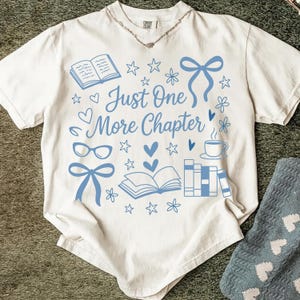以下が含まれることがあります： クリーム色のTシャツ。青色の「Just One More Chapter」の文字と、本と星のイラストが描かれています。Tシャツは丸首で半袖です。シルバーのネックレスが見えます。