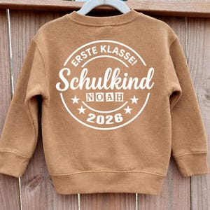 May include: A brown crewneck sweatshirt with white text. The text on the back reads "ERSTE KLASSE! Schulkind NOAH 2026" in a circular design. The sweatshirt is hanging on a white hanger against a wooden fence background.