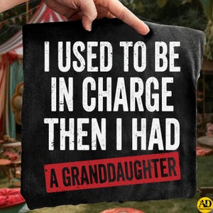 May include: Black t-shirt with white text that reads "I USED TO BE IN CHARGE THEN I HAD A GRANDDAUGHTER" in a distressed font. The text "A GRANDDAUGHTER" is in a red rectangle. The shirt is held up by a hand.