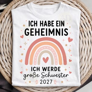 May include: A white t-shirt featuring a graphic with the text ICH HABE EIN GEHEIMNIS and ICH WERDE gro&szlig;e Schwester 2027 in black lettering. The design includes a multi-colored pastel rainbow, small pink hearts, and star shapes surrounding the text.