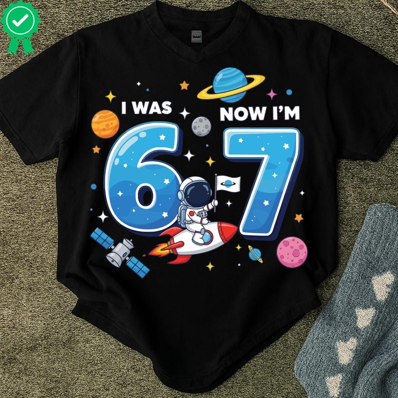 May include: Black v-neck t-shirt with a colourful space-themed graphic. The design features the text "I WAS 6 NOW I'M 7" with planets, stars, and an astronaut on a rocket ship. A fun birthday shirt.