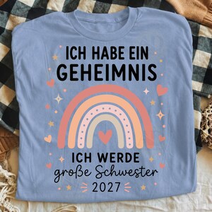 May include: A light blue t-shirt featuring a graphic of a multi-colored pastel rainbow with a heart in the center. The text reads: ICH HABE EIN GEHEIMNIS ICH WERDE gro&szlig;e Schwester 2027. The design includes small stars and hearts surrounding the rainbow.