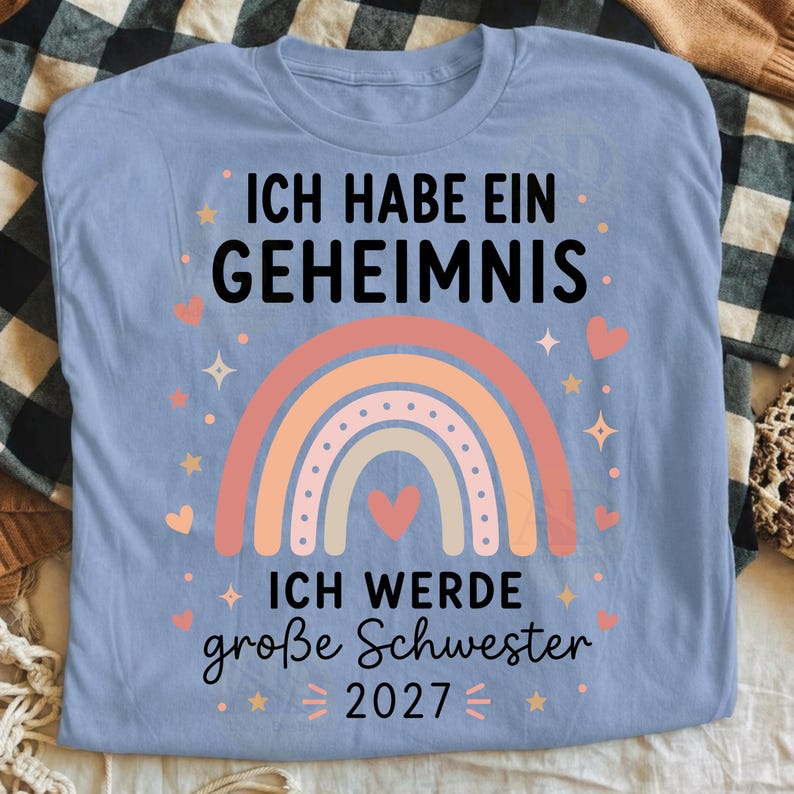 May include: A light blue t-shirt featuring a graphic of a multi-colored pastel rainbow with a heart in the center. The text reads: ICH HABE EIN GEHEIMNIS ICH WERDE gro&szlig;e Schwester 2027. The design includes small stars and hearts surrounding the rainbow.