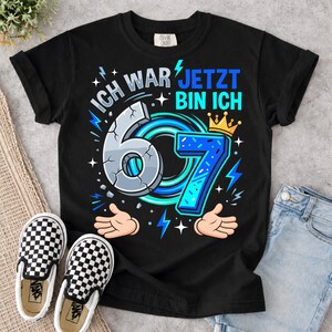May include: A black short-sleeve t-shirt featuring a graphic design with the text "ICH WAR 6 JETZT BIN ICH 7". The design includes a cracked number 6, a blue number 7 with a yellow crown, cartoon hands, and blue lightning bolt accents.