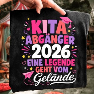 May include: A black t-shirt featuring colourful text that reads KITA ABGÄNGER 2026 EINE LEGENDE GEHT VOM GELÄNDE. The design includes a pink graduation cap, small stars, hearts, and a paper airplane graphic in shades of pink, purple, and yellow.