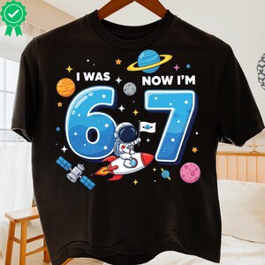 May include: Black t-shirt with a colourful space-themed graphic. The design features the text "I WAS 6 NOW I'M 7" with a cartoon astronaut on a rocket, planets, stars, and a satellite. A fun birthday shirt.