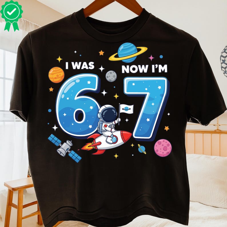 May include: Black t-shirt with a colourful space-themed graphic. The design features the text "I WAS 6 NOW I'M 7" with a cartoon astronaut on a rocket, planets, stars, and a satellite. A fun birthday shirt.