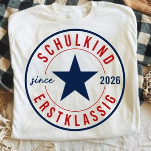 May include: A white short-sleeved t-shirt featuring a circular graphic with a dark blue star in the center. The text "SCHULKIND" is printed in red above the star, "ERSTKLASSIG" in red below, and "since 2026" in dark blue on the sides.