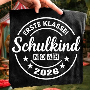 May include: Black t-shirt with white text. The shirt features the words "ERSTE KLASSE! Schulkind" in a circular design, with the name "NOAH" and the year "2026" below. The shirt is held up by a hand.