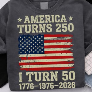 Puede incluir: Sudadera gris oscuro con el texto "AMERICA TURNS 250" y "I TURN 50" en una fuente blanquecina desgastada. El dise&ntilde;o incluye una bandera estadounidense desgastada. Las fechas 1776-1976-2026 est&aacute;n impresas debajo.
