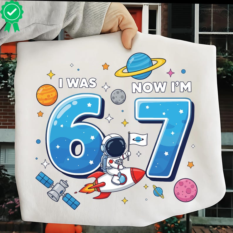 May include: White fabric featuring a space-themed design with the text "I WAS 6 NOW I'M 7" in blue. The design includes planets, stars, and an astronaut on a rocket. A fun birthday theme.