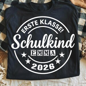 May include: Black t-shirt with a white circular design. The design includes the words "ERSTE KLASSE! Schulkind" and the name "EMMA" in block letters, along with the year "2026" and star accents. The shirt is laid on a plaid surface.