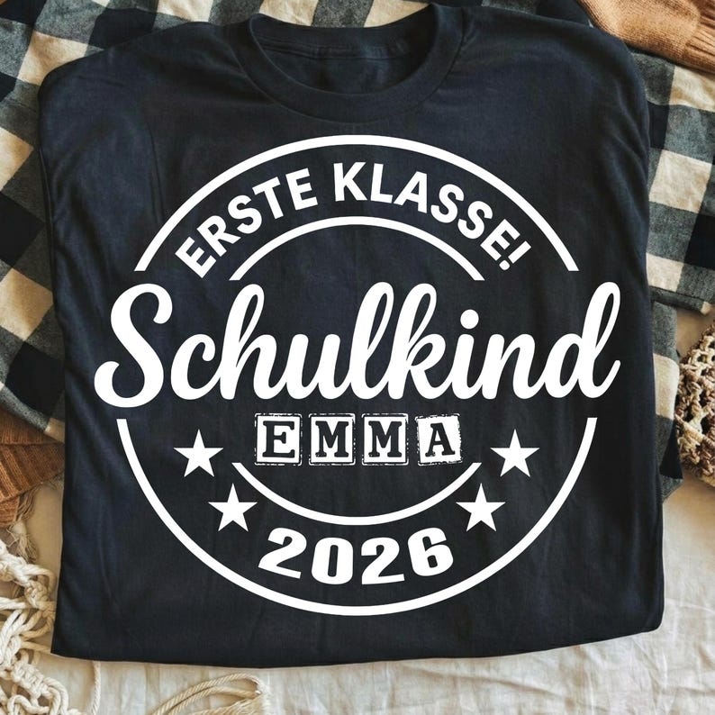 May include: Black t-shirt with a white circular design. The design includes the words "ERSTE KLASSE! Schulkind" and the name "EMMA" in block letters, along with the year "2026" and star accents. The shirt is laid on a plaid surface.