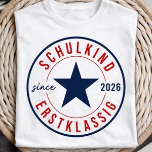 May include: A white short-sleeved t-shirt featuring a circular graphic with a dark blue star in the center. The text "SCHULKIND" is arched in red at the top, "ERSTKLASSIG" is arched in red at the bottom, and "since 2026" is in dark blue.