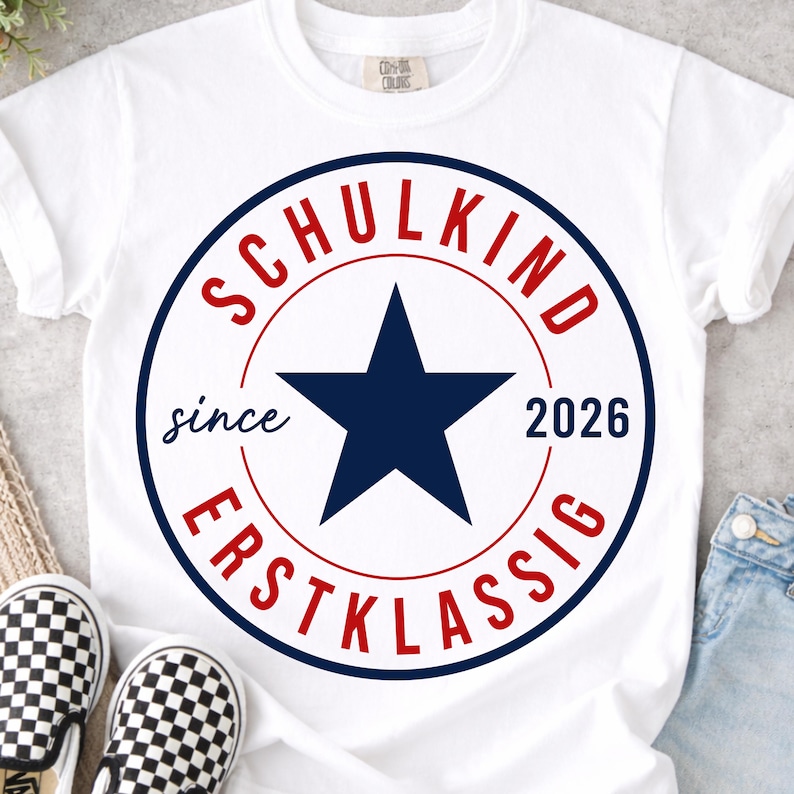 May include: A white short-sleeved t-shirt featuring a circular graphic with a dark blue star in the center. The text SCHULKIND is in red at the top, ERSTKLASSIG is in red at the bottom, and the words since 2026 are printed in dark blue.