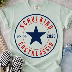 May include: A light green short-sleeve t-shirt featuring a circular graphic with a dark blue star in the center. The text reads SCHULKIND in red at the top, ERSTKLASSIG in red at the bottom, and since 2026 in dark blue on the sides.