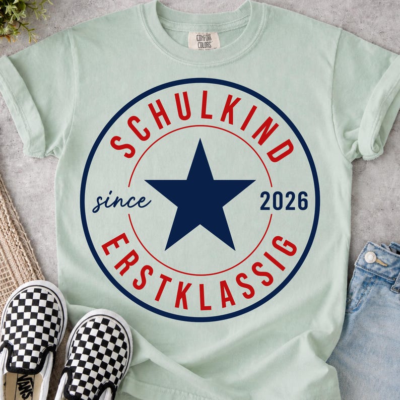 May include: A light green short-sleeve t-shirt featuring a circular graphic with a dark blue star in the center. The text reads SCHULKIND in red at the top, ERSTKLASSIG in red at the bottom, and since 2026 in dark blue on the sides.