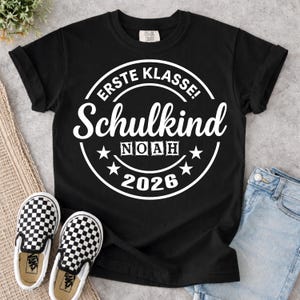 Könnte beinhalten: Schwarzes T-Shirt mit weißem, kreisförmigem Design, das "ERSTE KLASSE! Schulkind NOAH 2026" zeigt. Das Bild zeigt auch schwarz-weiß karierte Slip-On-Schuhe und blaue Jeans.
