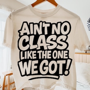 May include: Cream-colored, short-sleeved t-shirt with the phrase "AIN'T NO CLASS LIKE THE ONE WE GOT!" in bold, black lettering. The shirt is hanging on a wooden hanger against a soft, neutral background.