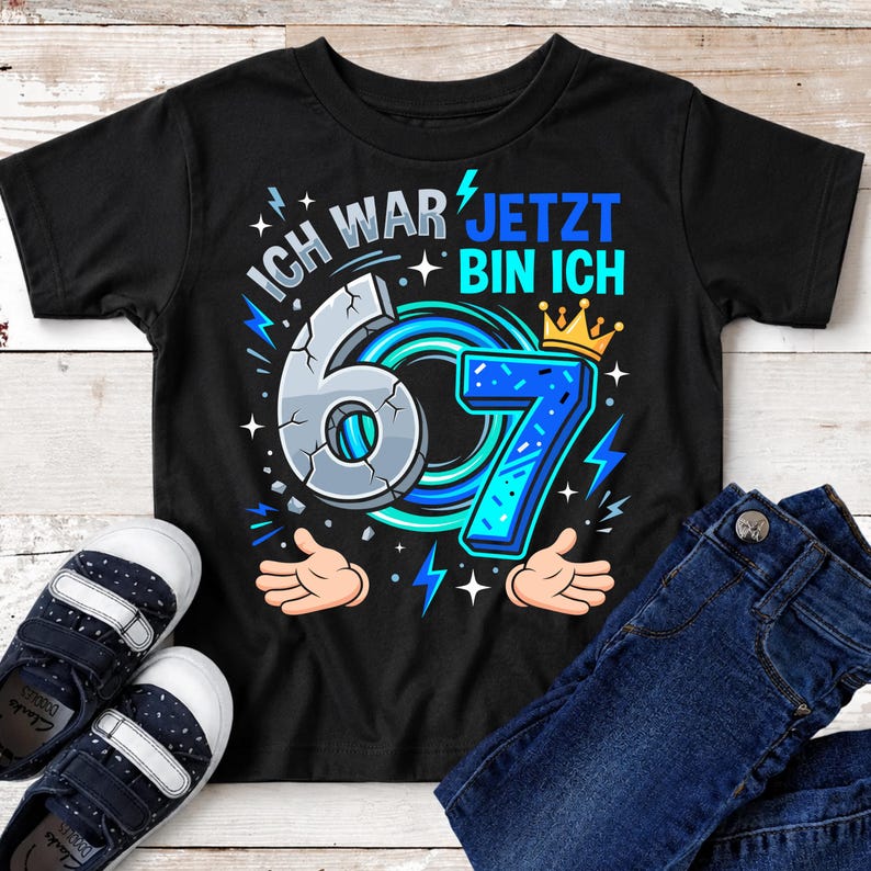 May include: A black short-sleeve t-shirt featuring a graphic with the text ICH WAR 6 JETZT BIN ICH 7. The design includes a cracked grey number 6, a blue number 7 with a yellow crown, blue lightning bolts, and two cartoon hands reaching upward.