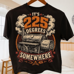 May include: A black short-sleeve t-shirt featuring a graphic of a barrel-style barbecue smoker with smoke rising from the chimney. The text reads IT'S 225 DEGREES SOMEWHERE in orange and white lettering, accompanied by a fork and spatula icon.