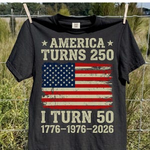 Puede incluir: Camiseta negra con el texto "AMERICA TURNS 250" y "I TURN 50" encima y debajo de una bandera americana desgastada. Las fechas 1776-1976-2026 est&aacute;n impresas debajo.