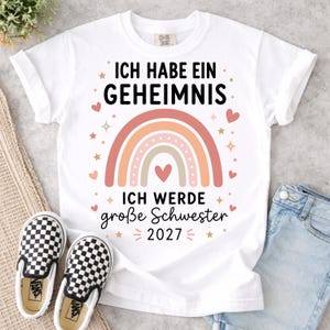 May include: A white short-sleeved t-shirt featuring a graphic of a multi-colored rainbow with a heart in the center. The text reads ICH HABE EIN GEHEIMNIS and ICH WERDE gro&szlig;e Schwester 2027, surrounded by small hearts and star icons in pink and gold.