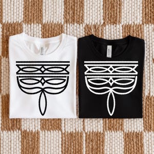 May include: Two t-shirts, one white and one black, each featuring a black and white graphic design. The design is a stylized mask or face, with beaded lines and diamond shapes. The shirts are laid flat on a patterned surface.