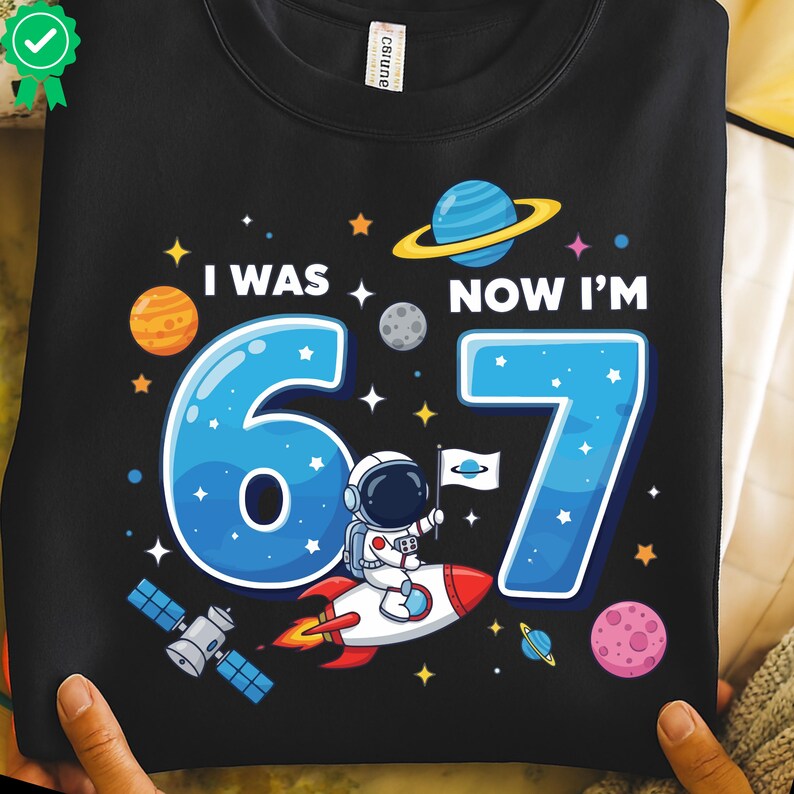 May include: Black sweatshirt featuring a space-themed design with the text "I WAS 6, NOW I'M 7". The graphic includes planets, stars, and an astronaut on a rocket. The numbers 6 and 7 are in blue.