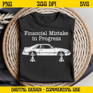 Könnte beinhalten: Schwarzes T-Shirt mit dem Text "Financial Mistake in Progress" und einer weißen Illustration eines Autos auf Wagenhebern. Das T-Shirt liegt auf einem geflochtenen Korb.