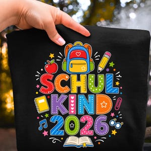 May include: A black t-shirt featuring a colorful graphic with the text SCHUL KIND 2026. The design includes a blue and yellow backpack, a red apple, a yellow pencil, a yellow notebook, an open book, and various small school-themed icons.