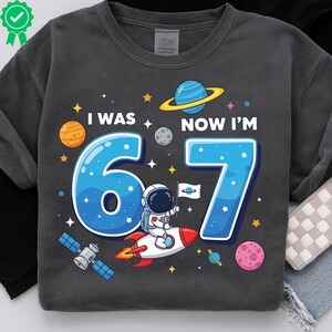 May include: Dark grey sweatshirt featuring a space-themed graphic. The design includes the text "I WAS 6, NOW I'M 7" in blue, along with planets, stars, and an astronaut on a rocket. A fun birthday shirt.