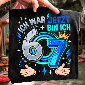 May include: A black t-shirt featuring a graphic with the text "ICH WAR JETZT BIN ICH" and the number "67". The number 6 is grey with a cracked texture, while the number 7 is blue with a gold crown on top, surrounded by blue lightning bolts.