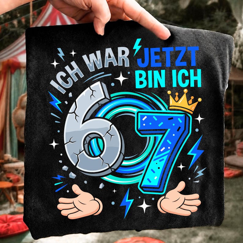 May include: A black t-shirt featuring a graphic with the text "ICH WAR JETZT BIN ICH" and the number "67". The number 6 is grey with a cracked texture, while the number 7 is blue with a gold crown on top, surrounded by blue lightning bolts.