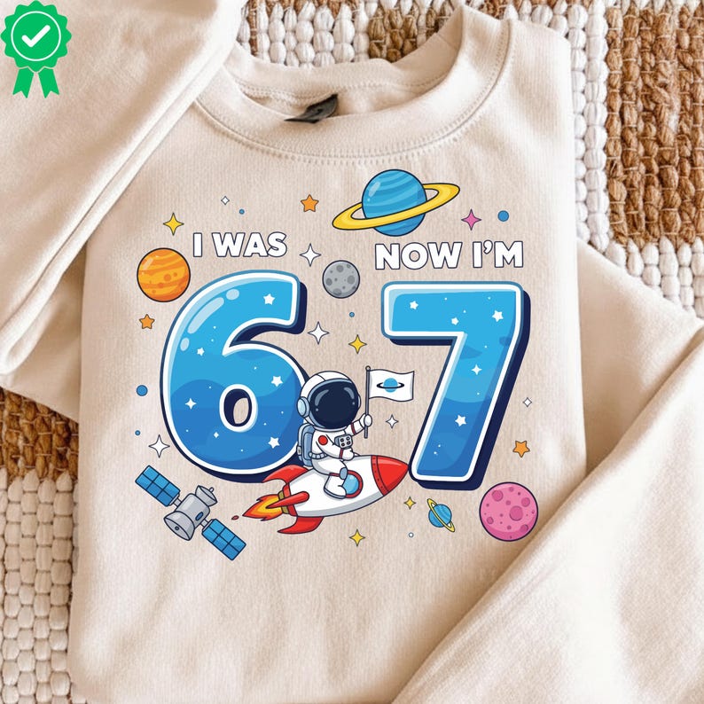 May include: A cream-coloured sweatshirt with a space-themed graphic. The graphic includes the text "I WAS 67 NOW I'M", blue numbers, an astronaut on a rocket, planets, and stars. The design is suitable for a 67th birthday.