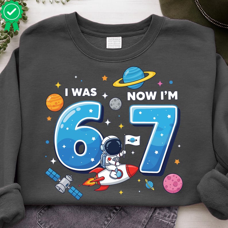 May include: Dark grey sweatshirt with a colourful space-themed graphic. The design features the text "I WAS 6, NOW I'M 7" with planets, stars, and an astronaut on a rocket. The numbers 6 and 7 are in blue.