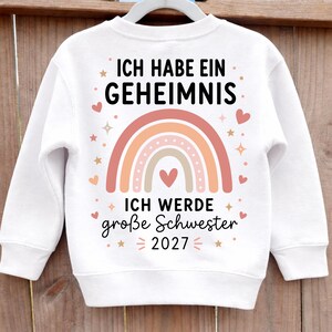 May include: A white long-sleeve sweatshirt featuring a central graphic of a muted-tone rainbow and hearts. The text reads: ICH HABE EIN GEHEIMNIS, ICH WERDE gro&szlig;e Schwester, and 2027, surrounded by small star and heart illustrations.