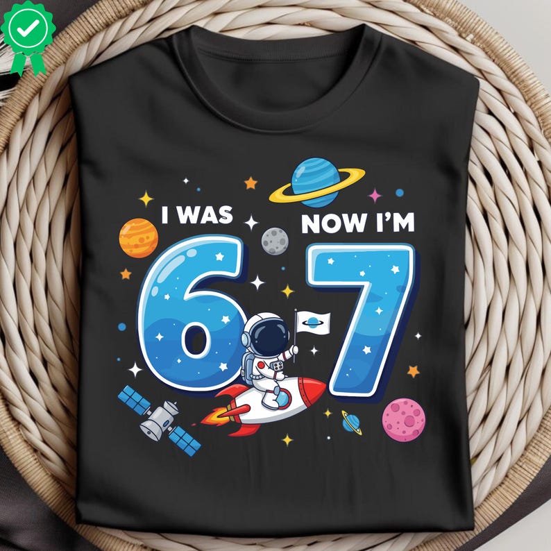 May include: Black t-shirt featuring a space-themed design with the text "I WAS 6, NOW I'M 7". The graphic includes planets, stars, and an astronaut on a rocket. A birthday shirt for a child.