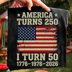 Puede incluir: Camiseta negra con el texto "AMERICA TURNS 250" y "I TURN 50". El dise&ntilde;o incluye una bandera estadounidense desgastada en rojo, blanco y azul. Las fechas 1776-1976-2026 est&aacute;n impresas debajo.
