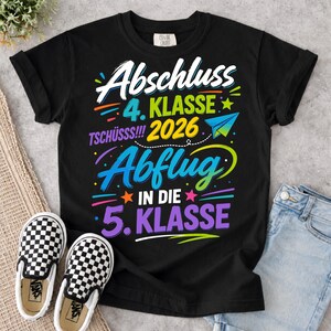 Könnte beinhalten: Schwarzes T-Shirt mit einem farbenfrohen Grafikdesign. Das Design enthält die Wörter "Abschluss 4. Klasse 2026 Tschüss!!! Abflug in die 5. Klasse". Ein Paar schwarz-weiß karierte Slip-On-Schuhe sind im Vordergrund.