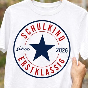 May include: A white t-shirt featuring a circular graphic with a dark blue star in the center. The text "SCHULKIND" is printed in red above the star, "ERSTKLASSIG" in red below, and "since 2026" in dark blue on the sides of the star.
