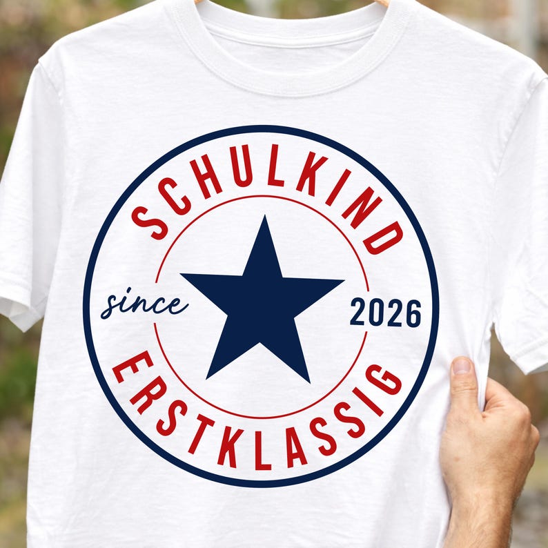 May include: A white t-shirt featuring a circular graphic with a dark blue star in the center. The text "SCHULKIND" is printed in red above the star, "ERSTKLASSIG" in red below, and "since 2026" in dark blue on the sides of the star.